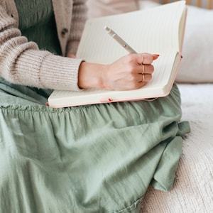 Puede incluir: Una persona escribe en un cuaderno con l&iacute;neas con un bol&iacute;grafo. El cuaderno tiene una cubierta rosa. La persona lleva un vestido verde claro y una rebeca beige. La escena est&aacute; ambientada en interiores, con un fondo suave de tonos neutros.