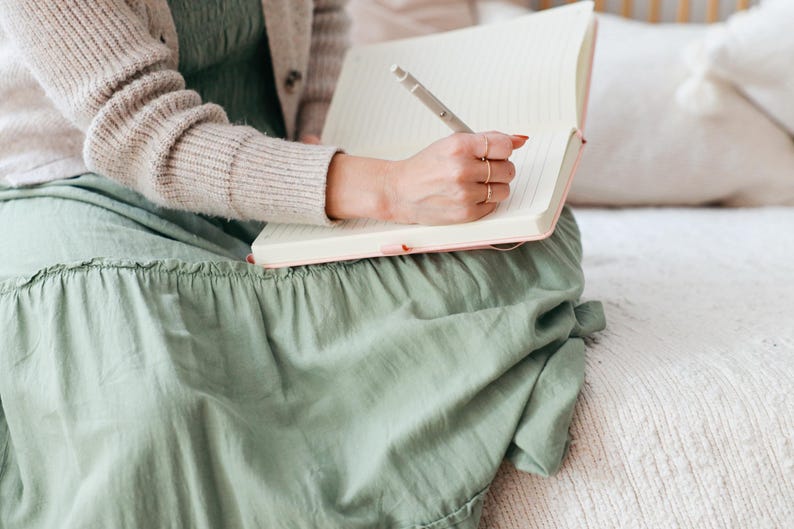 Puede incluir: Una persona escribe en un cuaderno con l&iacute;neas con un bol&iacute;grafo. El cuaderno tiene una cubierta rosa. La persona lleva un vestido verde claro y una rebeca beige. La escena est&aacute; ambientada en interiores, con un fondo suave de tonos neutros.