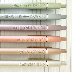May include: Six pastel-colored pens with silver accents and engraved text. The pens are arranged horizontally on lined paper. The phrases on the pens include "SMASH THE PATRIARCHY", "GIRLS WILL BE GIR POWERFUL", and "PEACE. LOVE. FEMINISM."