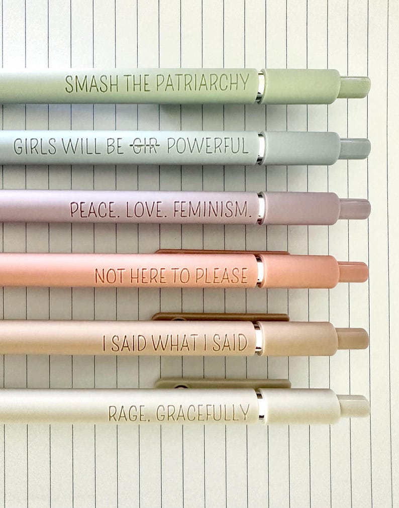 May include: Six pastel-colored pens with silver accents and engraved text. The pens are arranged horizontally on lined paper. The phrases on the pens include "SMASH THE PATRIARCHY", "GIRLS WILL BE GIR POWERFUL", and "PEACE. LOVE. FEMINISM."