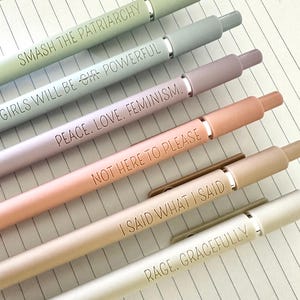 May include: Six pastel-colored pens with silver accents and matching caps. Each pen is engraved with a different empowering phrase: "SMASH THE PATRIARCHY," "GIRLS WILL BE GIR POWERFUL," "PEACE. LOVE. FEMINISM," "NOT HERE TO PLEASE," "I SAID WHAT I SAID," and "RAGE, GRACEFULLY."