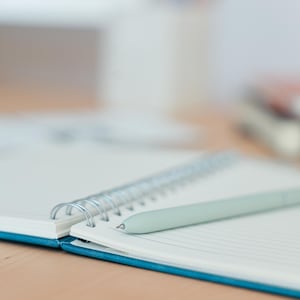 K&ouml;nnte beinhalten: Ein blaues Spiralblock mit einer silbernen Spiralbindung und einem hellgrauen Stift, der auf den ge&ouml;ffneten Seiten liegt.