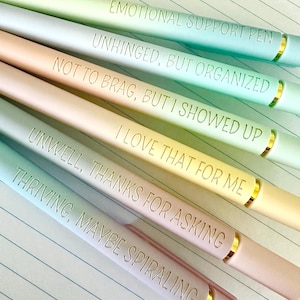Puede incluir: Un conjunto de seis bol&iacute;grafos de colores pastel con detalles dorados. Cada bol&iacute;grafo tiene una frase diferente grabada: "EMOTIONAL SUPPORT PEN", "UNHINGED, BUT ORGANIZED", "NOT TO BRAG, BUT I SHOWED UP", "I LOVE THAT FOR ME", "UNWELL, THANKS FOR ASKING", y "THRIVING, MAYBE SPIRALING".
