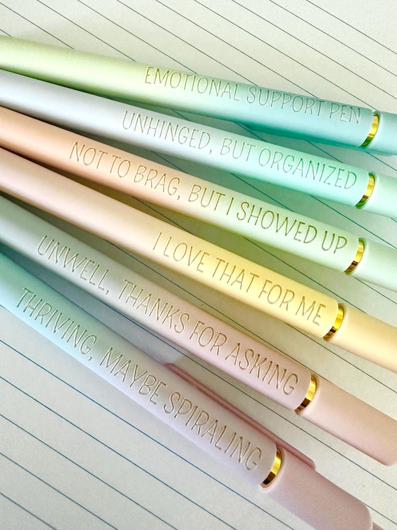 Puede incluir: Un conjunto de seis bol&iacute;grafos de colores pastel con detalles dorados. Cada bol&iacute;grafo tiene una frase diferente grabada: "EMOTIONAL SUPPORT PEN", "UNHINGED, BUT ORGANIZED", "NOT TO BRAG, BUT I SHOWED UP", "I LOVE THAT FOR ME", "UNWELL, THANKS FOR ASKING", y "THRIVING, MAYBE SPIRALING".