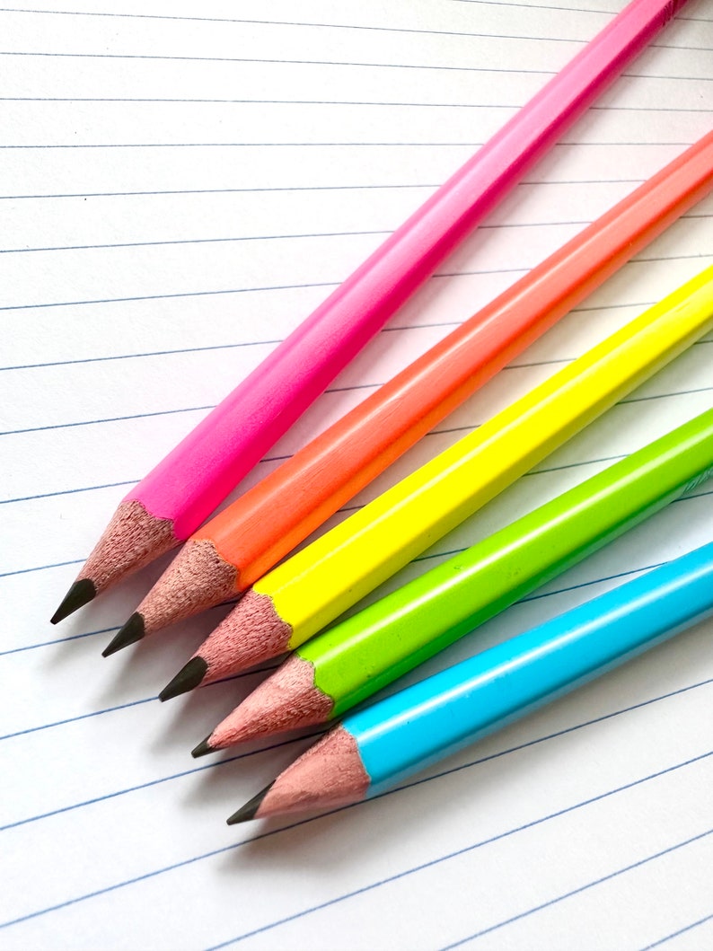 May include: Four colored pencils on a lined notebook. The pencils are pink, orange, yellow, and blue.