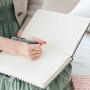 K&ouml;nnte beinhalten: Eine Person schreibt in ein graues, liniertes Notizbuch mit einem grauen Stift. Das Notizbuch ist ge&ouml;ffnet und die Seiten sind leer. Die Person tr&auml;gt ein gr&uuml;nes Kleid und einen beigefarbenen Cardigan.