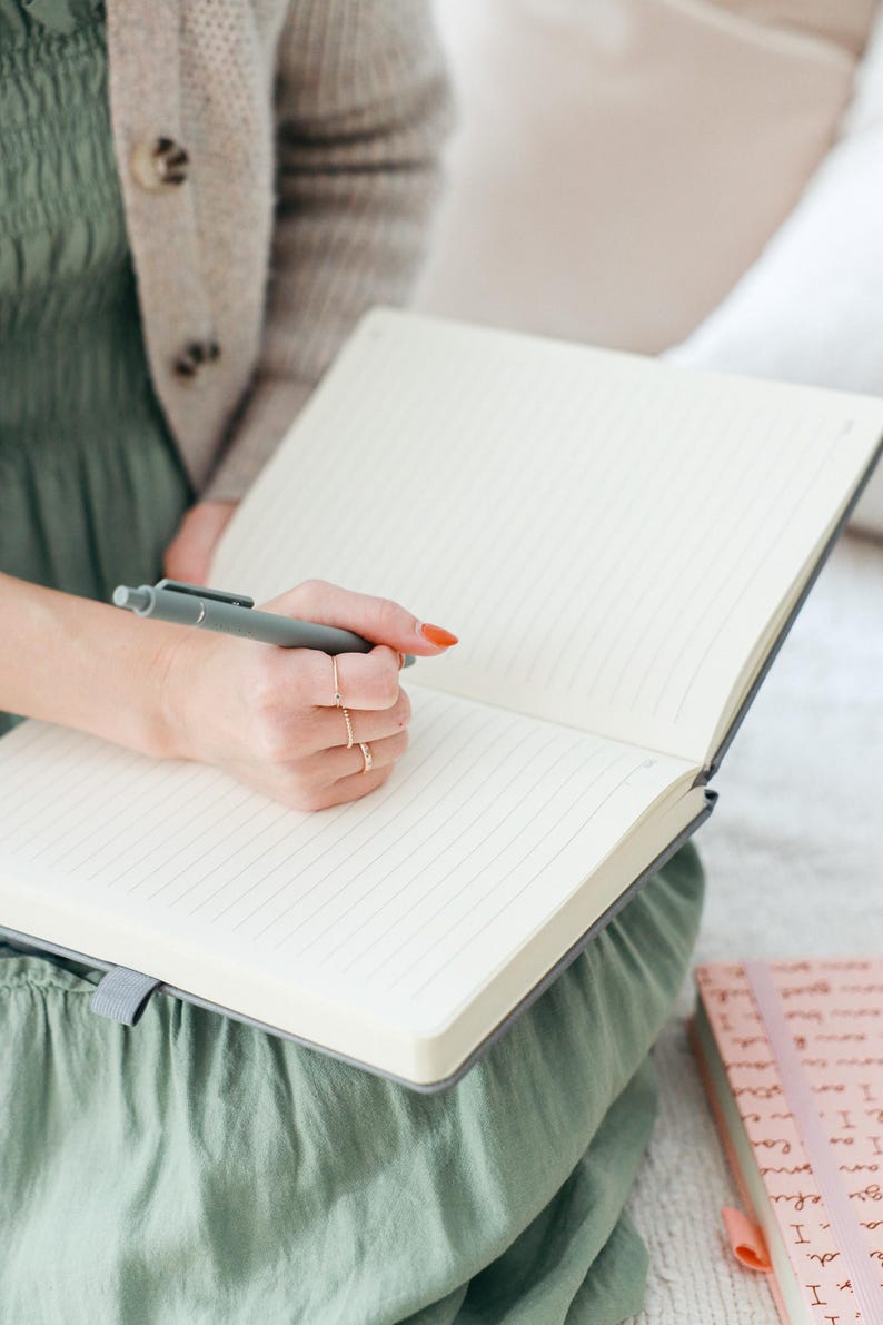 K&ouml;nnte beinhalten: Eine Person schreibt in ein graues, liniertes Notizbuch mit einem grauen Stift. Das Notizbuch ist ge&ouml;ffnet und die Seiten sind leer. Die Person tr&auml;gt ein gr&uuml;nes Kleid und einen beigefarbenen Cardigan.