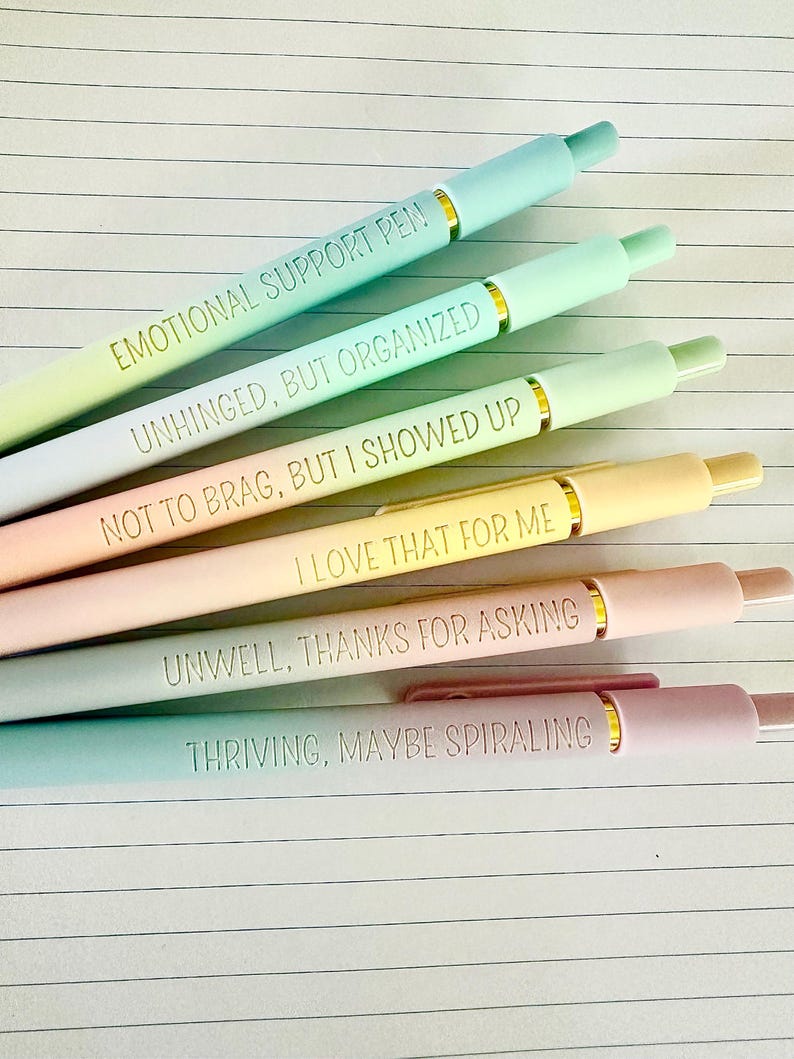 Puede incluir: Una colecci&oacute;n de bol&iacute;grafos de colores pastel con detalles dorados y frases humor&iacute;sticas. Los bol&iacute;grafos son en tonos azul, verde, amarillo, rosa y melocot&oacute;n. Cada bol&iacute;grafo tiene una frase diferente, incluyendo "EMOTIONAL SUPPORT PEN" y "THRIVING, MAYBE SPIRALING."