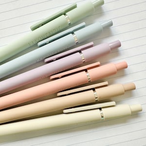 Puede incluir: Un surtido de bol&iacute;grafos de colores pastel, incluyendo tonos de verde, azul, rosa y beige, dispuestos diagonalmente sobre papel rayado. Cada bol&iacute;grafo tiene el nombre "Mackenzie" impreso en el cuerpo. Los bol&iacute;grafos tienen detalles plateados y clips a juego.