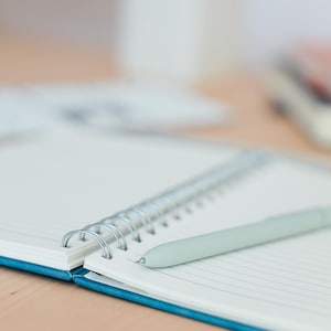 Puede incluir: Un cuaderno abierto, con espiral, con p&aacute;ginas rayadas y una cubierta turquesa. Un bol&iacute;grafo verde claro descansa sobre las p&aacute;ginas abiertas. El cuaderno est&aacute; sobre una superficie marr&oacute;n claro, con objetos borrosos en el fondo.