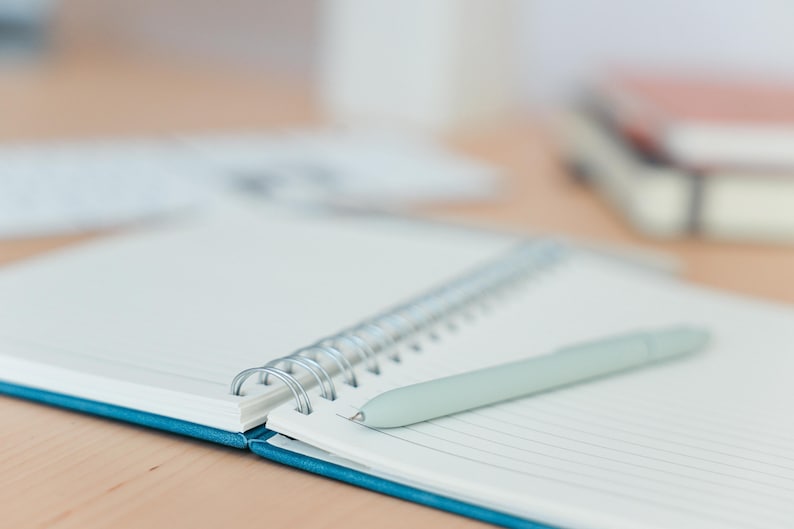 Puede incluir: Un cuaderno abierto, con espiral, con p&aacute;ginas rayadas y una cubierta turquesa. Un bol&iacute;grafo verde claro descansa sobre las p&aacute;ginas abiertas. El cuaderno est&aacute; sobre una superficie marr&oacute;n claro, con objetos borrosos en el fondo.