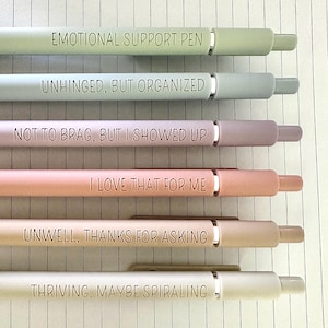 Puede incluir: Seis bol&iacute;grafos de colores pastel con detalles plateados y frases humor&iacute;sticas grabadas. Las frases incluyen "EMOTIONAL SUPPORT PEN", "UNHINGED, BUT ORGANIZED" y "THRIVING, MAYBE SPIRALING". Los bol&iacute;grafos est&aacute;n colocados sobre papel rayado.