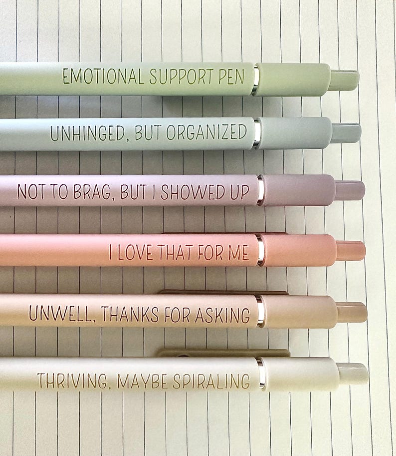 Puede incluir: Seis bol&iacute;grafos de colores pastel con detalles plateados y frases humor&iacute;sticas grabadas. Las frases incluyen "EMOTIONAL SUPPORT PEN", "UNHINGED, BUT ORGANIZED" y "THRIVING, MAYBE SPIRALING". Los bol&iacute;grafos est&aacute;n colocados sobre papel rayado.