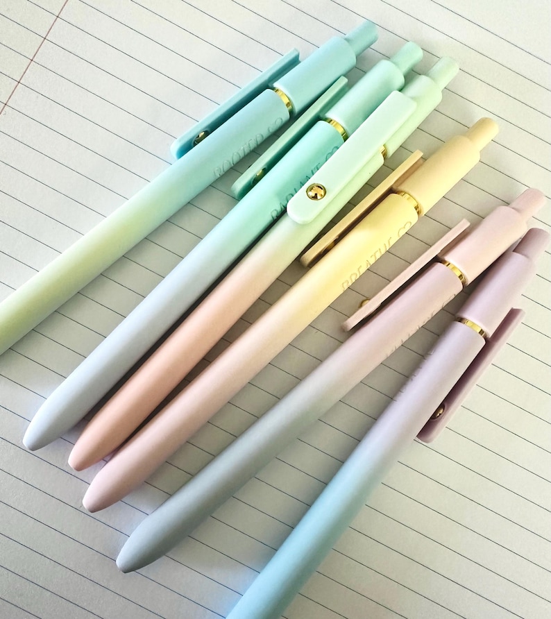 Puede incluir: Seis bol&iacute;grafos de colores pastel dispuestos en diagonal sobre papel rayado. Los bol&iacute;grafos presentan un degradado de color, que va del azul claro al rosa y al amarillo. Cada bol&iacute;grafo tiene un clip dorado y el texto "BREATHE CO" es visible en algunos clips.