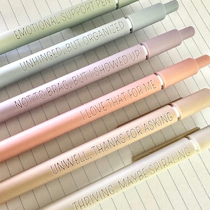 Puede incluir: Una colecci&oacute;n de seis bol&iacute;grafos de colores pastel con detalles plateados, cada uno con una frase humor&iacute;stica. Los bol&iacute;grafos est&aacute;n dispuestos en diagonal sobre papel rayado. Las frases incluyen "EMOTIONAL SUPPORT PEN" y "UNWELL, THANKS FOR ASKING."
