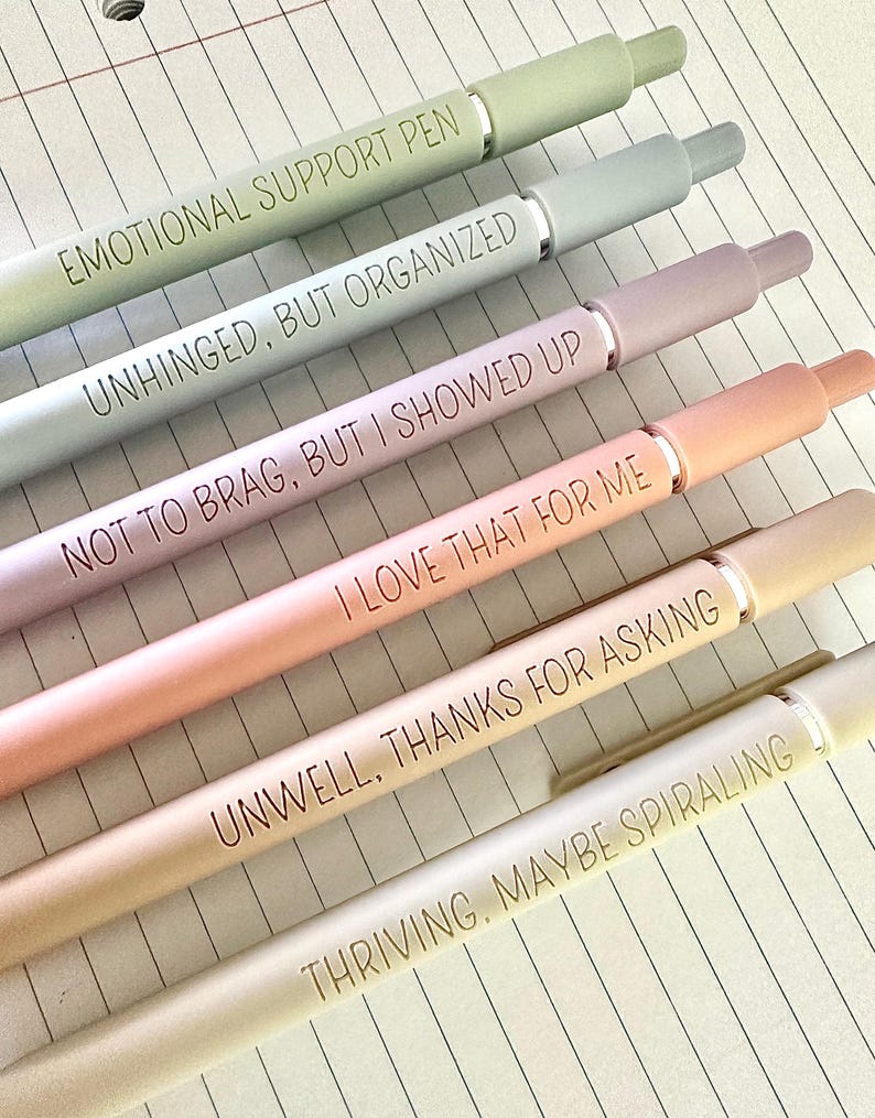 Puede incluir: Una colecci&oacute;n de seis bol&iacute;grafos de colores pastel con detalles plateados, cada uno con una frase humor&iacute;stica. Los bol&iacute;grafos est&aacute;n dispuestos en diagonal sobre papel rayado. Las frases incluyen "EMOTIONAL SUPPORT PEN" y "UNWELL, THANKS FOR ASKING."