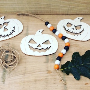 May include: Three wooden pumpkin ornaments with different carved faces, a spool of twine, and a Halloween-themed beaded garland with orange, black, and white beads.