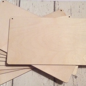 May include: A stack of eight unfinished wooden signs with rounded corners and holes for hanging. The signs are made of light-colored wood and are ready to be painted or stained.
