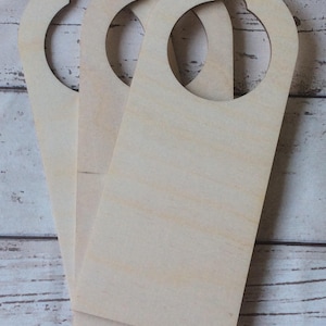 May include: Three unfinished wooden door hangers with a circular opening at the top and a rectangular body. The wood has a light, natural color and visible grain. The hangers are stacked slightly, showing their shape.