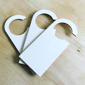 May include: Three blank white door hangers with a curved top and rectangular body are stacked on a light wood surface. The hangers are made of a smooth, solid material, and are ready for customisation.
