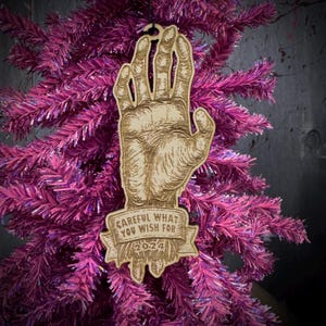 May include: Wooden Christmas ornament featuring a detailed hand illustration. The ornament has the text "CAREFUL WHAT YOU WISH FOR 2024" on a banner. The ornament is hanging on a pink Christmas tree.