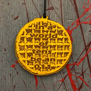 May include: A yellow, waffle-shaped ornament with the text "In a world of tens, be an 011." The ornament is hanging against a gray background with red paint splatters. The text and waffle pattern are in a darker brown.