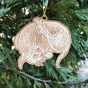 May include: A wooden Christmas ornament in the shape of a cat, hanging from a green string. The ornament is light brown with etched details, depicting the cat's fur and features. The ornament is hanging in front of a green evergreen tree.