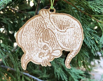 Könnte beinhalten: Ein hölzerner Weihnachtsschmuck in Form einer Katze, der an einer grünen Schnur hängt. Der hellbraune Schmuck weist geätzte Details auf, die das Fell und die Merkmale der Katze darstellen. Er hängt vor einem Hintergrund aus grünem Nadelbaumlaub.