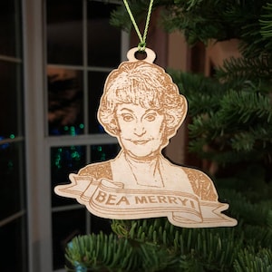 May include: Wooden Christmas ornament featuring a portrait of a woman with the text "BEA MERRY!" on a banner. The ornament is hanging on a Christmas tree. The portrait is etched into the wood, and the ornament has a green string for hanging.