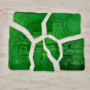 Pode incluir: Uma obra de arte abstrata verde sobre uma superfície de madeira clara. A obra é composta por várias formas fragmentadas e irregulares que se encaixam, criando um design coeso. As formas verdes têm uma aparência texturizada, sugerindo uma impressão ou pintura.