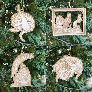 May include: Four wooden cat-themed Christmas ornaments hanging on a green tree. The ornaments feature different cat poses, including a cat grooming itself, a cat in a frame, and a cat on a rock. Each ornament is made of light-colored wood.