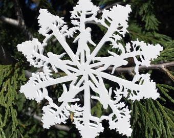 May include: White snowflake ornament with a unique design. The ornament features a detailed cutout of a person walking through a forest scene. The snowflake is hanging against a backdrop of green foliage.