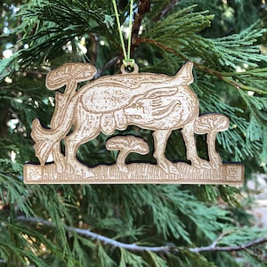 May include: A wooden Christmas ornament featuring a stylized deer with mushrooms. The ornament is light brown with a dark border and a green string for hanging. The ornament is set against a backdrop of green pine needles.