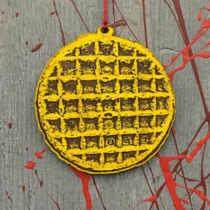 May include: Yellow wooden waffle ornament with a grid pattern and a red string for hanging. The ornament has a circular shape and is painted with yellow and brown colors. The background is gray with red paint splatters.