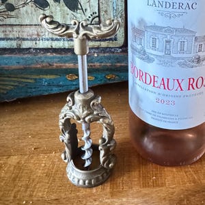 May include: A bottle of Bordeaux Rosé wine with a label that reads "CHEVALIER de LANDERAC BORDEAUX ROSE 2023". A decorative brass corkscrew is next to the bottle. The wine bottle is a light pink colour.
