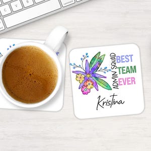 May include: A white coffee mug with a brown liquid sits on a square coaster. A second coaster features a floral design with the text "BEST TEAM EVER" and the name "Kristina". A computer keyboard is in the background.