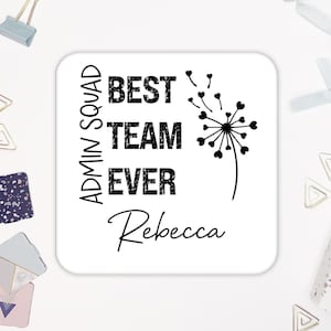 May include: A white square coaster with the text "ADMIN SQUAD BEST TEAM EVER Rebecca" in black. A black dandelion graphic is on the right side. Office supplies, including paper clips and binder clips, are in the background.