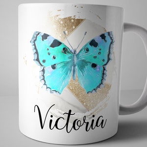 May include: White ceramic mug with a blue butterfly graphic and gold glitter accents. The mug has the name "Victoria" written in black cursive lettering.
