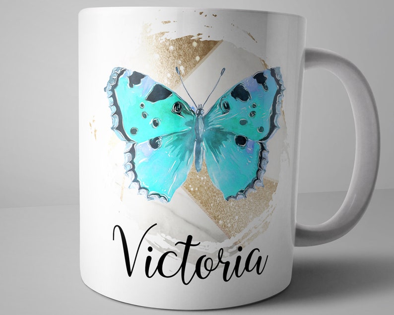 May include: White ceramic mug with a blue butterfly graphic and gold glitter accents. The mug has the name "Victoria" written in black cursive lettering.
