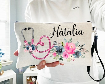 May include: A white canvas makeup bag with a black zipper and a black strap. The bag has a pink stethoscope design with a floral pattern and the name "Natalia" printed in black.