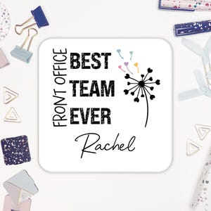 May include: White square coaster with black text that reads "Front Office Best Team Ever" and a black dandelion with colorful hearts. The name "Rachel" is written in cursive below the dandelion.