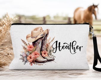 May include: A white zippered pouch featuring a printed illustration of a tan cowboy hat resting on a pair of patterned cowboy boots, surrounded by peach-colored flowers and wheat stalks. The name Heather is printed in black script on the right.