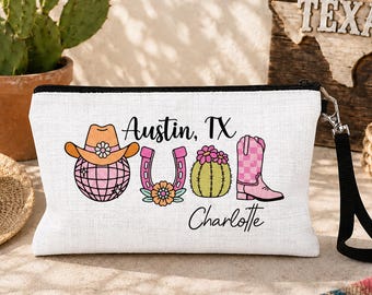 May include: A white rectangular zippered pouch with a black wrist strap featuring a graphic print of a cowboy hat on a disco ball, a horseshoe, a cactus with flowers, and a pink checkered cowboy boot. The text reads Austin, TX and Charlotte.
