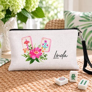 May include: A rectangular off-white zippered pouch featuring a printed design of two mahjong tiles, a vibrant pink flower, and green foliage. The name Linda is printed in black script on the right side. A black wrist strap is attached to the side.
