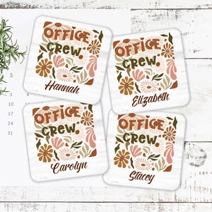 May include: Four square coasters with a floral design and the words "OFFICE CREW" in brown and green. Each coaster has a different name: Hannah, Elizabeth, Carolyn, and Stacey. The background is white with a wood grain pattern.