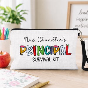 May include: A white rectangular zippered pouch with a black wrist strap. The front features the black text Mrs. Chandler's and SURVIVAL KIT, with the word PRINCIPAL in large, colorful, patterned letters designed to look like school supplies.