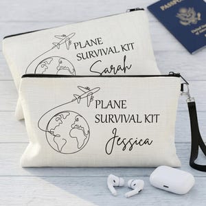 May include: Two off-white Plane Survival Kit pouches with black zips and wrist straps. Each pouch features a black line drawing of a plane flying around the globe, with the text "PLANE SURVIVAL KIT" and a name. A passport, earbuds, and charging case are visible.