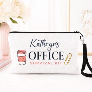 May include: A white rectangular zippered pouch with a black wrist strap. The front features the text Kathryn's OFFICE SURVIVAL KIT in black and pink, accompanied by a graphic of a pink coffee cup with a heart and a gold paperclip.