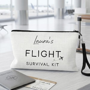 May include: A white rectangular zippered pouch with a black wrist strap. The front features the black printed text "Laura's FLIGHT SURVIVAL KIT" with a small airplane icon and a dashed flight path line trailing behind the airplane graphic.