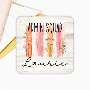 May include: Square coaster with a white wood grain background. The words "ADMIN SQUAD" are in black block letters above the cursive name "Laurie". Vertical brush strokes in orange and pink are decorated with floral illustrations.